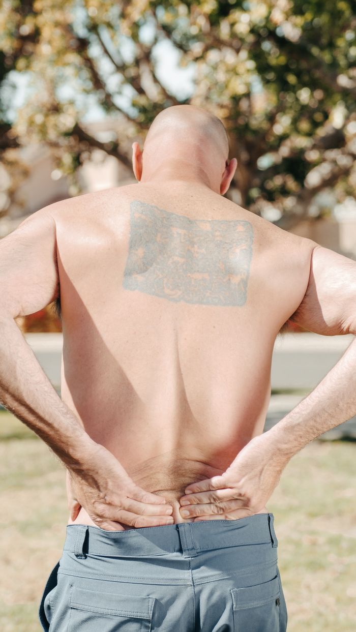 Shirtless bald man from behind holding his lower back, outdoors in sunny weather, with a large tattoo.