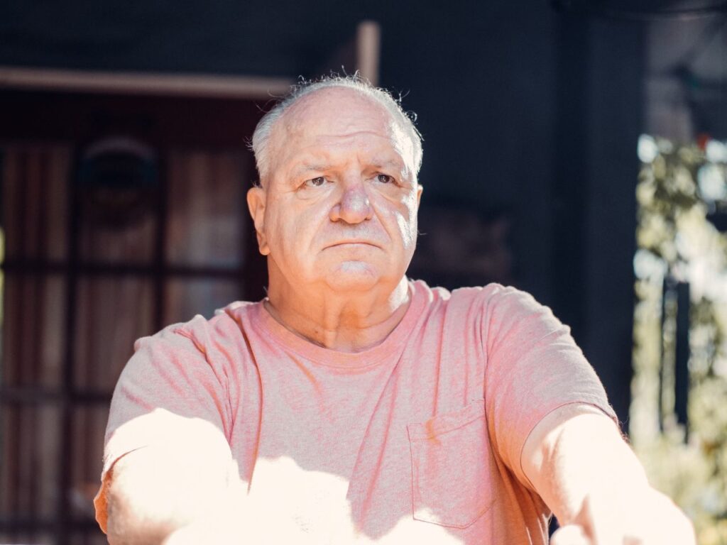 A senior man with a thoughtful expression, sitting outdoors in sunlight.